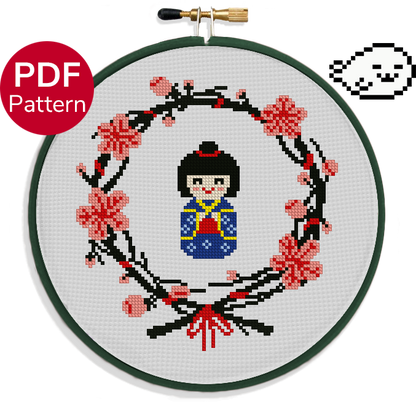 cross stitch pattern of a japanese wooden doll wearing a blue kimono surrounded by a wreath of sakura leaves